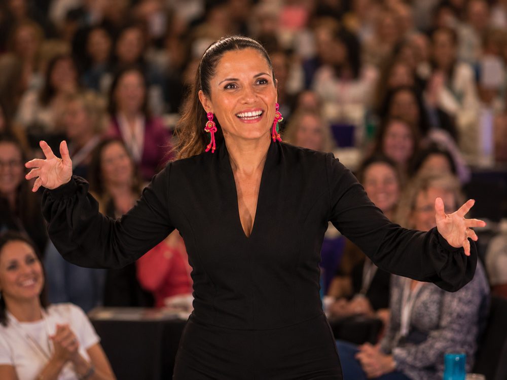 Melanie Schambach on stage with her arms open, speaking to a full auditorium