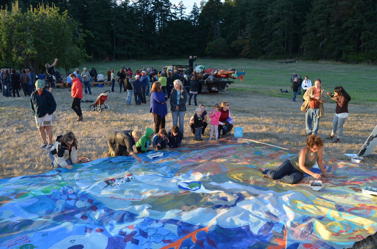 The travelling mural spread flat on the grass at a totem pole journey stop, people kneeling along its edges to paint while others gather behind